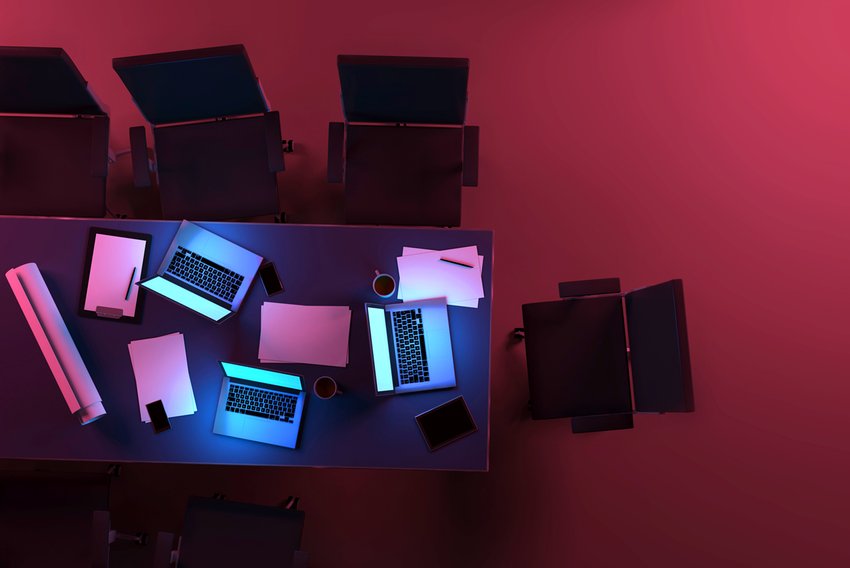 Three active laptop computers on a desk inside an office space at night, with glowing blue light from monitors illuminating the table. Aerial view, as seen from above. Purple light, in contrast with blue displays, illuminates the dark background. Teamwork and deadlines. Tablets, smartphones, coffee mugs, paper documents and projects are also visible on the desk, surrounded by office chairs. Dark purple and red hues at midnight hour. Copy space on dark areas. Digitally generated image.